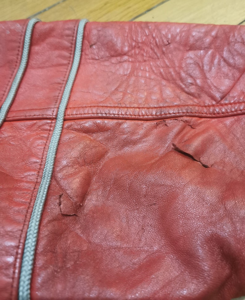May include: Close-up of a red leather bag with a white zipper. The leather has a textured surface and shows signs of wear.
