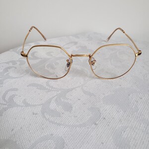 Pu&ograve; includere: Occhiali dorati con montatura ottagonale. Le montature sono sottili e delicate, con lenti trasparenti. Gli occhiali sono appoggiati su uno sfondo bianco a motivi, creando un'estetica pulita ed elegante.