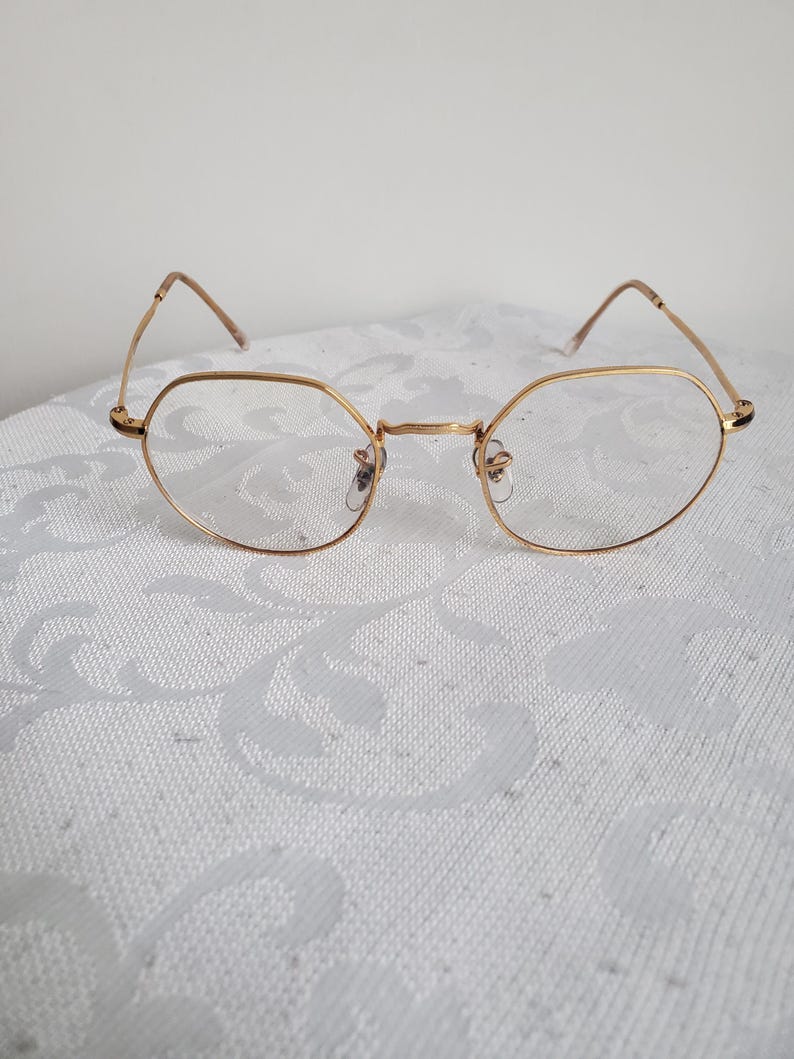 Pu&ograve; includere: Occhiali dorati con montatura ottagonale. Le montature sono sottili e delicate, con lenti trasparenti. Gli occhiali sono appoggiati su uno sfondo bianco a motivi, creando un'estetica pulita ed elegante.