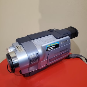 Peut inclure: Caméscope Sony Digital 8 argenté et noir avec zoom 700x. Le caméscope dispose des fonctions USB Streaming, SteadyShot et NightShot. Une sangle grise est attachée sur le côté.