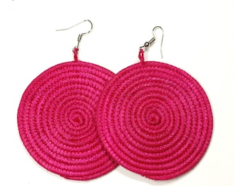 May include: A pair of large, round, pink earrings made of woven straw. The earrings have a spiral design and are attached to silver hooks.