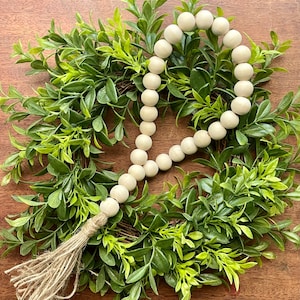 May include: A white wooden bead garland with a natural jute tassel. The garland is draped over a green leafy wreath.