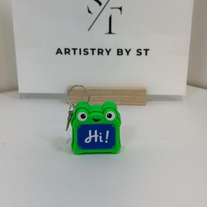 May include: A bright green frog-shaped keychain featuring a blue rectangular center with the word Hi! written in white script. The keychain includes a silver metal ring attached to the top, and the frog has two black eyes and a small smile.