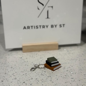 May include: A miniature keychain featuring a stack of three books in brown, black, and green. The keychain has a silver clasp and ring. In the background is a white sign with the text "ARTISTRY BY ST".