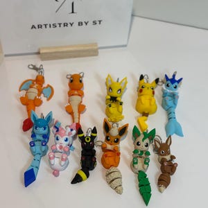 May include: A collection of colorful Pokémon character keychains. The keychains feature various Pokémon designs in orange, yellow, blue, pink, black, and brown. The keychains are attached to silver metal rings.