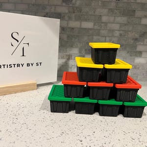 May include: A stack of small, black plastic storage containers with colorful lids in yellow, red, and green. The containers are arranged on a speckled surface. A white sign with the text "ARTISTRY BY ST" is in the background.