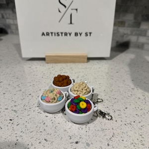 May include: Four miniature white ceramic bowls filled with colorful cereal, each attached to a keychain. The cereal includes chocolate puffs, frosted flakes, and colorful fruit loops. A sign in the background reads "ARTISTRY BY ST."