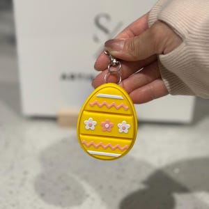 May include: Yellow Easter egg keychain with white and pink floral and zig-zag designs. The egg-shaped charm has a silver-colored clasp. A fun and festive accessory for Easter.