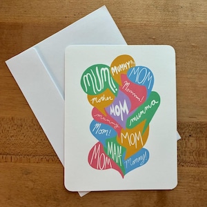 May include: A white greeting card with a colorful design and a white envelope. The card features the words "MUM," "MOM," "Mummy," and "Mommy" in various colors and fonts, arranged in speech bubbles.
