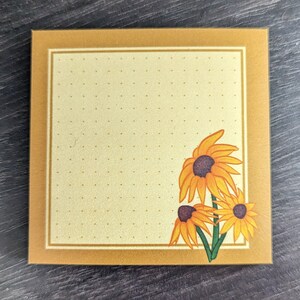 May include: Square notepad with a gold border and a light yellow interior dotted with small dots. The lower right corner features a cluster of three orange and yellow sunflowers with green stems.