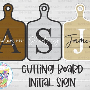 May include: Three wooden cutting boards with the letters A, S, and J on them. The boards are brown, gray, and beige. The text "Anderson", "Sherling", and "James" is written below the letters. The text "CUTTING BOARD INITIAL SIGN" is written below the boards.