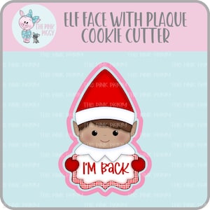 May include: A cookie cutter in the shape of an elf face with a red hat and a white collar. The elf holds a sign that says "I'M BACK" in red text. The design includes a pink and white gingham border.