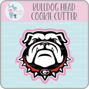 Puede incluir: Un cortador de galletas de cabeza de bulldog en blanco y negro con un collar rojo y una 'G' roja y blanca en el collar. El cortador de galletas está delineado en rosa.