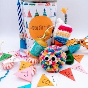 May include: A birthday party favor gift box with a colorful pom-pom, a party popper, a cake, a candle, and a banner that says "Happy Birthday".