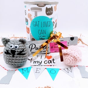 May include: A gift basket for cat lovers with a can that says "Cat Lover Can", a gray crocheted cat toy, a pink crocheted cat toy, a clear keychain with the text "I love my cat", a bag of cat treats, and a blue and white banner that says "Meow".