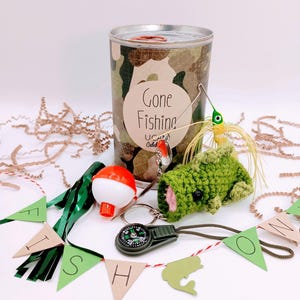 May include: A fishing-themed gift set with a camouflage-patterned container labeled "Gone Fishing." Includes a bobber, a compass, a crocheted fish, and a banner spelling "FISH." Decorative accents include green and brown streamers.