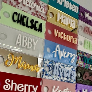May include: A collection of colorful acrylic name tags with various names written in white, black, and gold lettering. The tags are decorated with glitter and have a hole punched at the top for hanging.