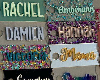May include: A collection of colorful, glittery keychains with various names, including Victoria, Rachel, Damien, and Emma. Each keychain features a unique design and color scheme.