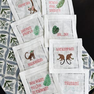 May include: A collection of white linen cocktail napkins with embroidered designs and text. Each napkin features a different design, including monkeys, leaves, and phrases like "Piper nol" and "Tsunami". The embroidery is in various colors.