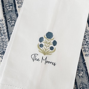 May include: White folded kitchen towel with a floral design in blue and green. The design features a cluster of stylized flowers and leaves, with the text "The Moores" embroidered below. The towel is on a blue and white patterned surface.