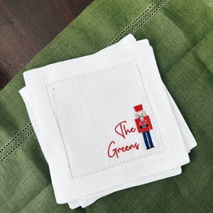 May include: White linen cocktail napkins with a red embroidered nutcracker and the text "The Greens." The napkins are stacked on a green linen placemat. The nutcracker is wearing a red coat and hat.