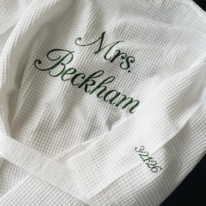 May include: White waffle-weave robe with a matching belt. The robe features green embroidered text that reads "Mrs. Beckham" and the number "32126" on the sleeve. The robe is folded, showing the textured fabric.