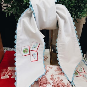 May include: White scarf with light blue scalloped edges and embroidered game card designs. The scarf is draped over a gold stand, with red and white patterned textiles in the background. The scarf features a unique, decorative design.