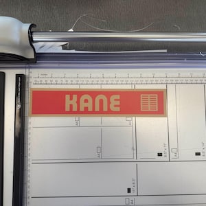 May include: A paper cutter with a white and black design. The cutter has a silver blade and a red label with the name "KANE" in beige letters. The cutting surface has measurements in inches.
