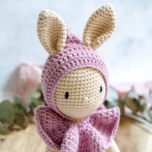 May include: A crocheted bunny toy wearing a pink hooded cape. The bunny has a cream-colored body and pink ears. The cape has a ruffled edge.