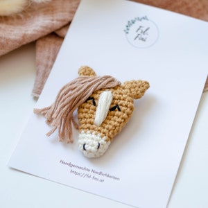 May include: A crocheted horse head pin with a brown body and a white face. The horse has a brown mane and a white stripe down its nose. The pin is handmade and is available for purchase on the website https://fr-fini.at.