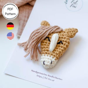 Könnte beinhalten: Ein gehäkelter Pferdekopf mit braunem Körper, weißer Schnauze und brauner Mähne. Das Pferd hat geschlossene Augen und ein kleines Lächeln. Das Pferd befindet sich auf einem weißen Hintergrund mit dem Text "Handgemachte Niedlichkeiten https://fri-fini.at".