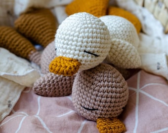 May include: Three crocheted stuffed duck toys in white, brown, and yellow. The ducks are lying on a pink and white patterned fabric.