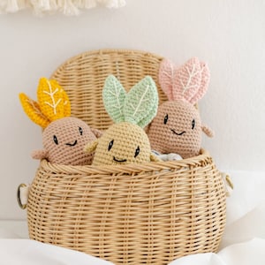 Könnte beinhalten: Drei gehäkelte Plüschtiere in Form von Gemüse in einem Weidenkorb. Die Spielzeuge sind hellbraun, gelb und rosa mit grünen Blättern. Jedes Spielzeug hat ein lächelndes Gesicht.