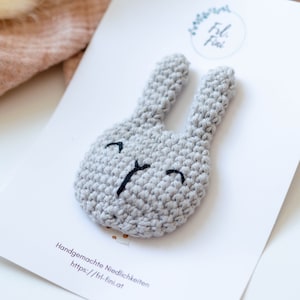 May include: A gray crocheted bunny with black embroidered eyes and a smiling mouth. The bunny is on a white card with the text "Handgemachte Niedlichkeiten https://frl-fini.at" printed on it.
