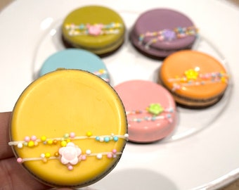 Puede incluir: Seis galletas decoradas con glaseado de colores pastel y chispas. Las galletas están dispuestas en un plato blanco. La galleta en primer plano es amarilla con una flor rosa y chispas blancas.