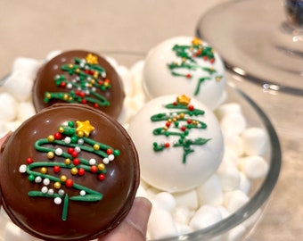 May include: Four chocolate and white chocolate Christmas tree hot cocoa bombs decorated with green, red, and white sprinkles. The bombs are sitting on a bed of white marshmallows.