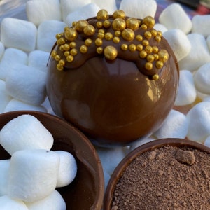 May include: A chocolate sphere covered in gold sprinkles, surrounded by white marshmallows. The chocolate sphere is sitting on a brown surface, next to a bowl of brown powder and a bowl of white marshmallows.