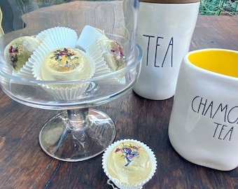 May include: A glass pedestal bowl filled with four white paper cupcake liners, each containing a clear, round, golden-coloured bath bomb with dried flowers inside. The bowl is on a wooden table next to a white ceramic tea canister with the word "TEA" written on it and a white ceramic mug with the words "CHAMOMILE TEA" written on it.