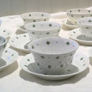 May include: A set of ten white teacups and saucers with a green floral pattern. The cups and saucers are arranged in a circle on a white tablecloth.