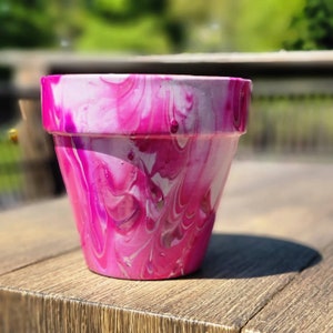 Puede incluir: Una maceta de terracota marmoleada en rosa y blanco. La maceta tiene un diseño de espiral con pintura rosa y blanca.
