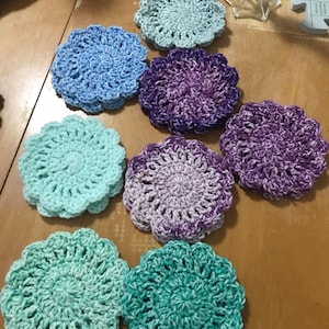 Puede incluir: Un conjunto de posavasos de ganchillo hechos a mano en varios colores, incluyendo azul claro, morado y turquesa. Los posavasos tienen forma circular con un borde festoneado y están colocados sobre una superficie de madera.
