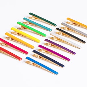May include: A set of 12 colorful hair clips with gold metal clasps. The clips are arranged in a row, with the colors ranging from green to purple.