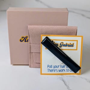 Peut inclure: Une pince &agrave; cheveux rectangulaire noire, emball&eacute;e dans une bo&icirc;te rose avec une pochette en velours rose. L'emballage comprend une carte jaune avec le texte "Pull your hair back. There's work to do." et le nom "Ann Gabriel" en lettres bleues et jaunes.