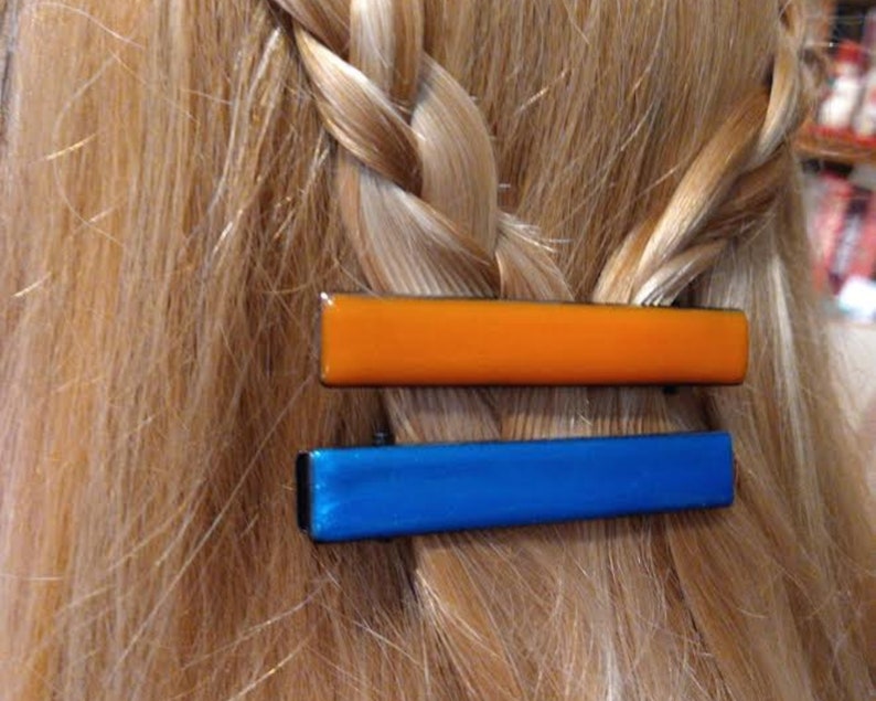Peut inclure: Deux pinces &agrave; cheveux, l'une orange et l'autre bleue, sont fix&eacute;es dans une tresse de cheveux blonds.