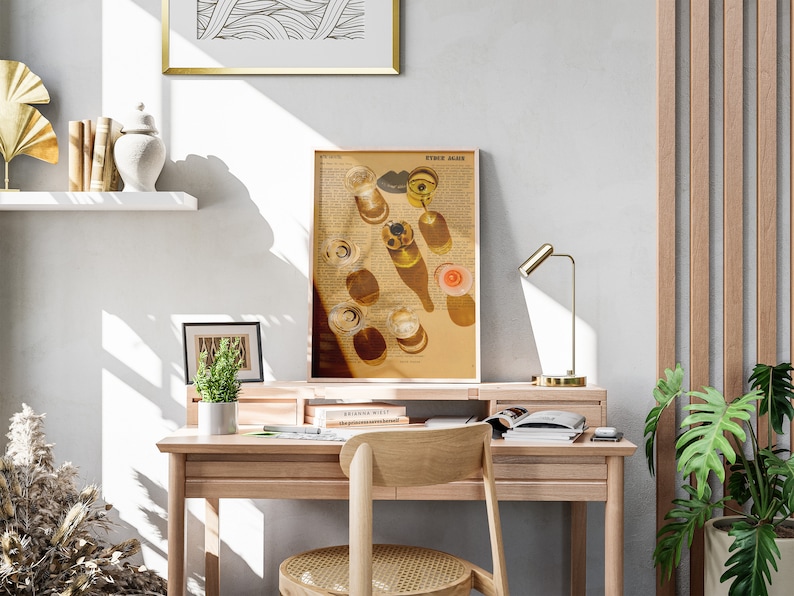 May include: A light wood desk with a gold lamp, a framed print of glasses of wine, and a potted plant. The desk has a drawer and a shelf. There are books on the shelf and a chair with a woven seat.