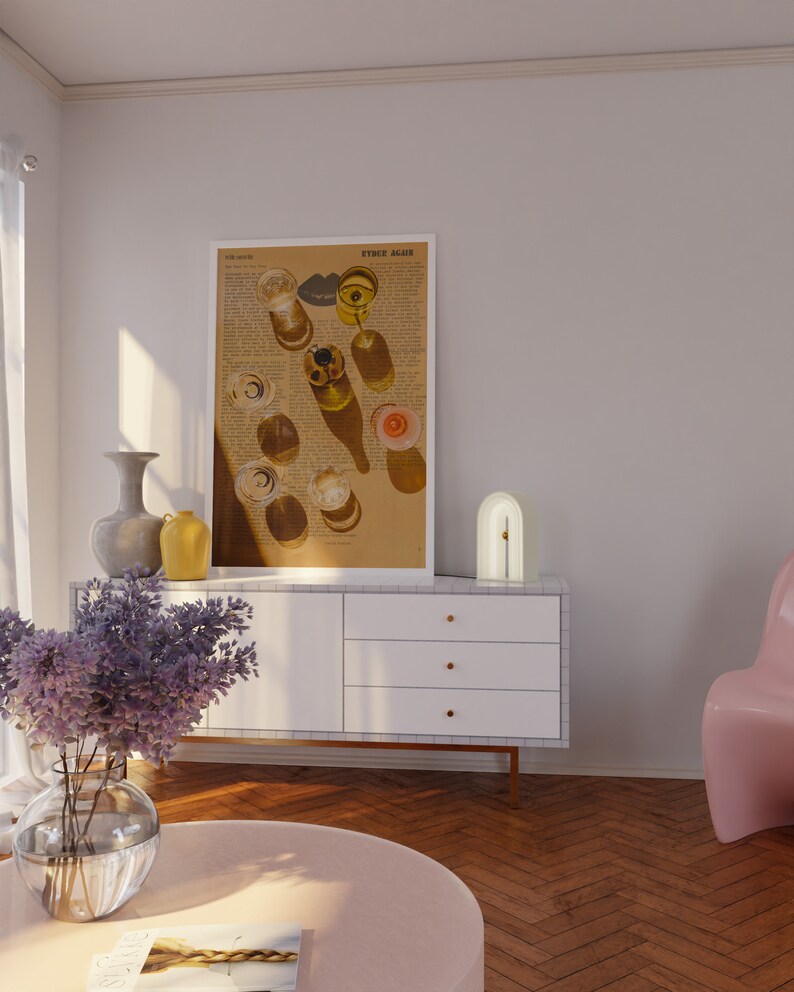 May include: A white cabinet with three drawers and gold handles sits on a wood floor. A white lamp with a gold arch sits on top of the cabinet. A vase with purple flowers sits on a pink round table in front of the cabinet. A framed print with the text "Either Again" hangs on the wall behind the cabinet.