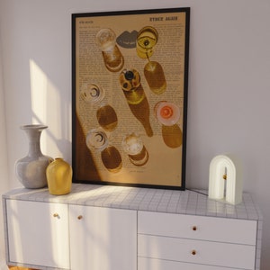 May include: A framed print with a black frame and a white background. The print features a photograph of wine glasses and bottles on a page of text. The text reads "RYDER AGAIN". The print is hanging on a white wall above a white cabinet with gold handles. A white arch-shaped lamp is on the cabinet.