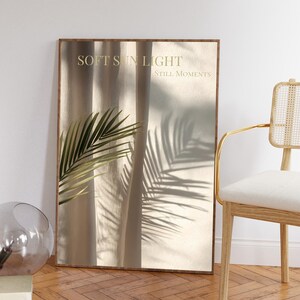 May include: A framed art print featuring the text "SOFT SUN LIGHT STILL MOMENTS." The artwork displays a palm leaf and its shadow on a beige curtain. A chair with a white cushion and gold frame is visible.