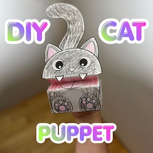 May include: A paper cat puppet featuring a gray colored head with large eyes, pink inner ears, and a curved tail. The mouth area is colored red with white teeth, and the base shows gray paws with pink pads. The text DIY CAT PUPPET is visible.