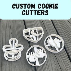 Puede incluir: Cortadores de galletas de plástico blanco con forma de chupete, pañal, huellas de pies de bebé y huellas de manos de bebé. El texto "CUSTOM COOKIE CUTTERS" está encima.